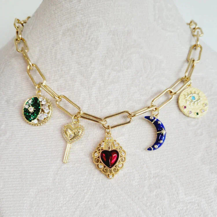 Close-up of a gold charm necklace featuring colorful pendants including a red heart, moon, key, and celestial symbols, displayed on a white textured background