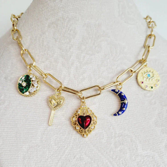 Close-up of a gold charm necklace featuring colorful pendants including a red heart, moon, key, and celestial symbols, displayed on a white textured background